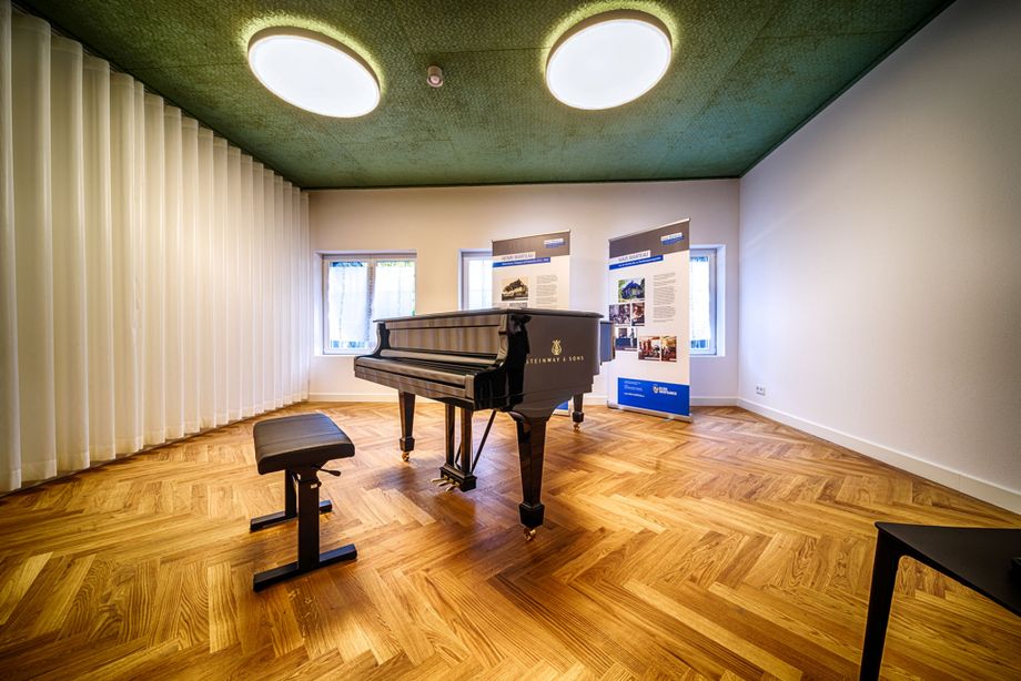 Ein Klavier steht in einem Raum, der mit Parkettboden ausgelegt ist. Der Raum wirkt edel.