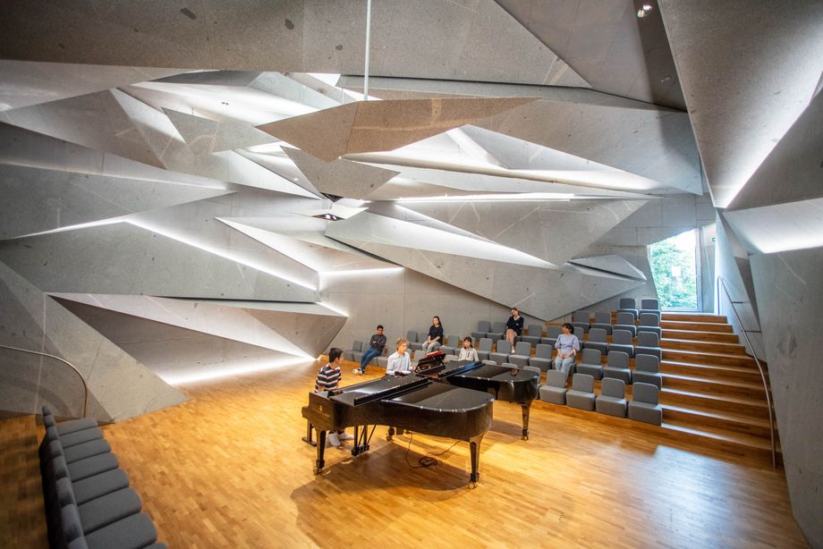 Zwei Personen sitzen in einem futuristisch gestalteten Raum an zwei Konzertflügeln. Einige Personen hören zu.