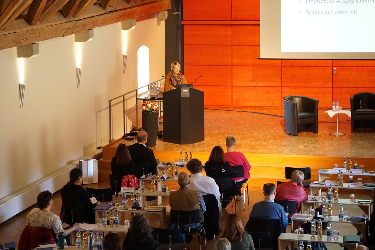 Blick in den Tagungsraum, Rednerin an schwarzem Pult hält Präsentation vor Publikum